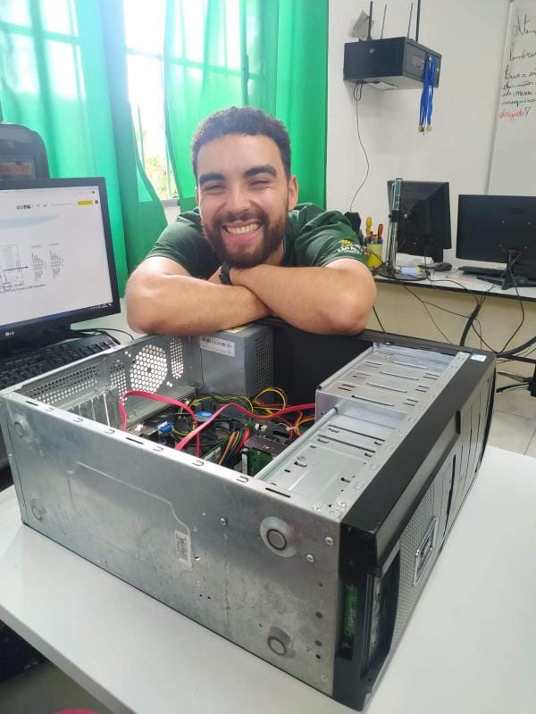 Um homem branco, com barba e cabelos castanhos, vestido de camiseta verde escuro está sorrindo, com braços cruzados em cima de um computador preto aberto. Ao fundo, há uma janela com cortinas verdes, paredes brancas e outros 3 computadores pretos. 